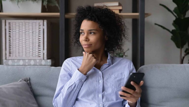 A woman holding her phone and looking out her window while thinking