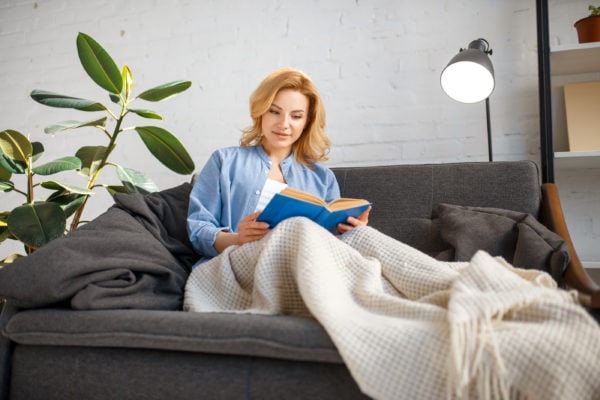 Young woman under a blanket reading book on couch